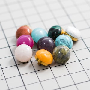 May include: A collection of ten small, round, dome-shaped knobs with smooth, polished surfaces in various colors including pink, orange, purple, turquoise, white, green, and brown, each featuring a gold-toned metal base with a threaded screw.