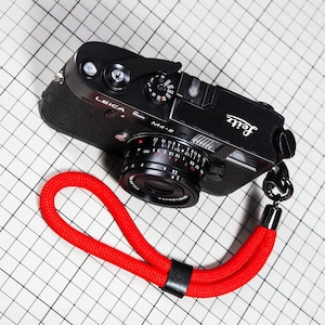 May include: A black Leica M4-2 camera with a red wrist strap. The camera has a black lens and silver accents. The red wrist strap is made of a woven material and has a black leather band. The camera is on a white grid background.