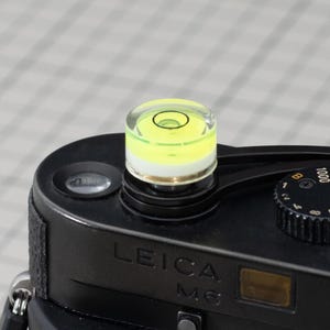 Puede incluir: Una cámara Leica M6 negra con un nivel de burbuja adjunto en la parte superior. El nivel tiene una parte superior transparente y un líquido amarillo en el interior. La cámara tiene una correa negra y un botón plateado. El texto de la cámara es visible.