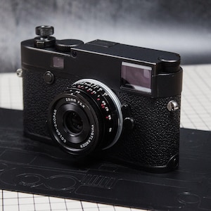 Puede incluir: Una cámara telemétrica negra con una lente de 28 mm, apoyada sobre una superficie negra. La cámara tiene un cuerpo texturizado y un anillo de lente plateado. La lente tiene marcas rojas y blancas. El fondo es gris borroso.