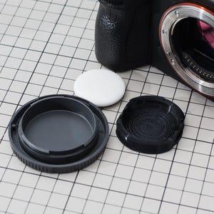 May include: A black digital camera with a lens mount, a lens cap, and a white circular object. The lens cap is black and has a ridged design. The camera body is black and has a textured grip.