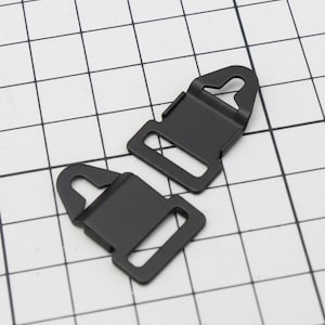 Puede incluir: Dos hebillas de metal negro con un marco rectangular y un gancho curvo. Las hebillas son de metal negro mate y están sobre un fondo de cuadrícula blanca.