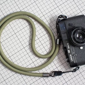 May include: A black Leica M4-2 camera with a lens attached, connected to a thick, olive green rope camera strap. The strap features metallic silver hardware at the connection points, and the camera body displays the text LEICA and M4-2.