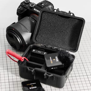 May include: A black Sony camera with a lens is positioned behind a black case containing audio equipment. The case holds a Rode Wireless GO II microphone and other accessories. A red coiled cable is visible.