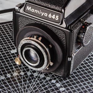 Op de afbeelding: Een zwarte Mamiya 645 middenformaat filmcamera met een getextureerde behuizing, een opvallende lens met zilveren metalen ringen en een sluitertijdknop aan de zijkant, geplaatst op een zwarte snijmat met witte rasterlijnen en markeringen.