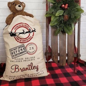 May include: A white canvas Santa sack with red and black lettering. The sack has a red and black stamp that says "List 2X Checked". The sack is decorated with a red and black illustration of Santa's sleigh and reindeer. The text on the sack reads "Express Delivery From The North Pole", "Do Not Open Before 25 December", "Please Deliver Enclosed Presents To Brantley", and "If Undeliverable Return To Santa Claus North Pole".