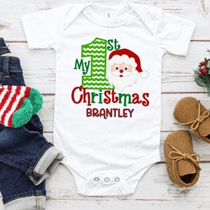 May include: White baby onesie with "My 1st Christmas" and a Santa Claus graphic. The name "Brantley" is below the text. The image also includes a pair of blue jeans, brown baby shoes, and a candy cane.