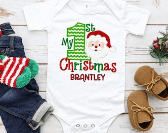 May include: White baby onesie with "My 1st Christmas" and a Santa Claus graphic. The name "Brantley" is below the text. The image also includes a pair of blue jeans, brown baby shoes, and a candy cane.