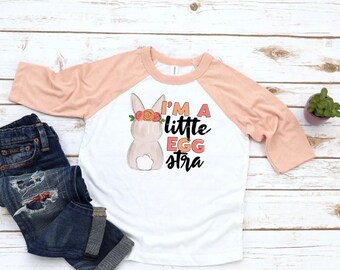 May include: A white and peach baseball tee shirt with a pink bunny rabbit wearing a flower crown and the text "I'm a little egg-stra".