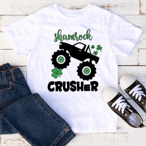 Pu&ograve; includere: Maglietta bianca con una grafica di un monster truck nero e il testo "shamrock crusher" in nero. Il camion ha disegni di trifoglio verde sui lati.
