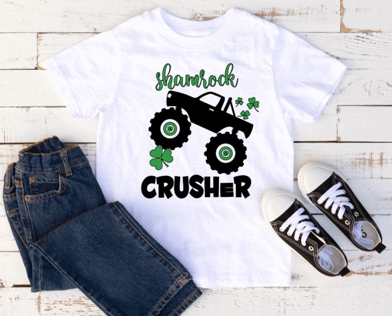 Pu&ograve; includere: Maglietta bianca con una grafica di un monster truck nero e il testo "shamrock crusher" in nero. Il camion ha disegni di trifoglio verde sui lati.