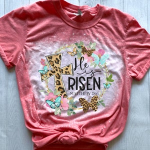 May include: A pink bleached t-shirt with a floral wreath design featuring a leopard print cross, butterflies, and the text "He is Risen Matthew 28:6".