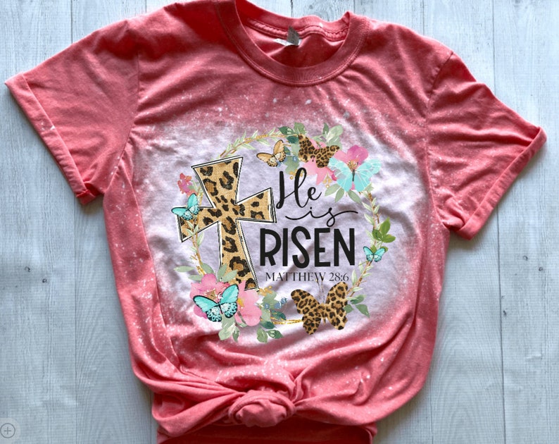May include: A pink bleached t-shirt with a floral wreath design featuring a leopard print cross, butterflies, and the text "He is Risen Matthew 28:6".