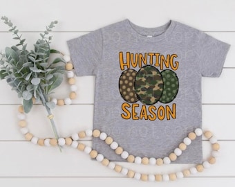 May include: A gray short-sleeved t-shirt with the text "Hunting Season" in orange. Three circles with camouflage and polka dot patterns are behind the text.