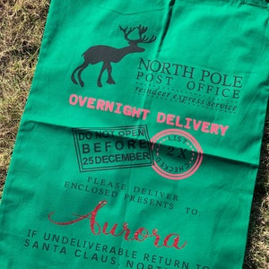 May include: A green canvas Santa sack with a reindeer graphic, the text "NORTH POLE POST OFFICE reindeer express service OVERNIGHT DELIVERY", and a pink stamp that says "LIST 2X CHECKED". The sack also has the text "DO NOT OPEN BEFORE 25 DECEMBER PLEASE DELIVER ENCLOSED PRESENTS TO: Aurora IF UNDELIVERABLE RETURN TO: SANTA CLAUS, NORTH POLE".
