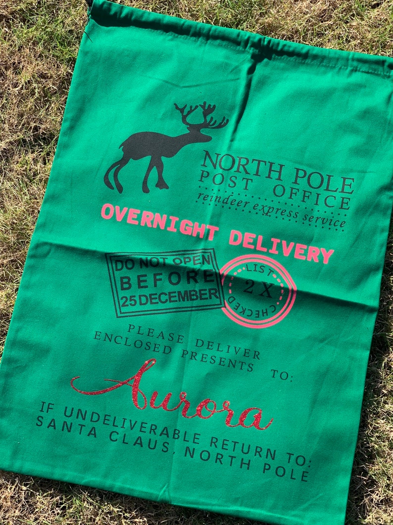 May include: A green canvas Santa sack with a reindeer graphic, the text "NORTH POLE POST OFFICE reindeer express service OVERNIGHT DELIVERY", and a pink stamp that says "LIST 2X CHECKED". The sack also has the text "DO NOT OPEN BEFORE 25 DECEMBER PLEASE DELIVER ENCLOSED PRESENTS TO: Aurora IF UNDELIVERABLE RETURN TO: SANTA CLAUS, NORTH POLE".