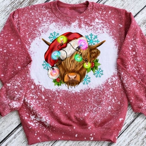 May include: Red sweatshirt with a white bleach design of a Highland cow wearing a Santa hat and Christmas lights.