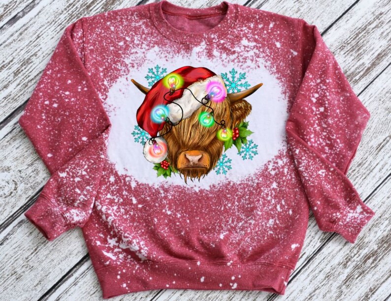 May include: Red sweatshirt with a white bleach design of a Highland cow wearing a Santa hat and Christmas lights.