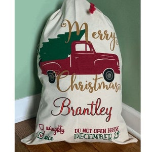 May include: A white Christmas sack with a red truck carrying a green Christmas tree. The sack has the text "Merry Christmas Brantley" in gold lettering. The sack also has a checklist with the options "naughty" and "nice" and the text "Do Not Open Before December 25".