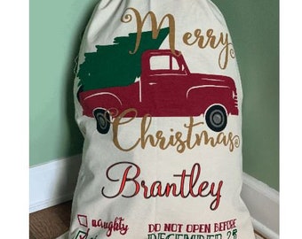 May include: A white Christmas sack with a red truck carrying a green Christmas tree. The sack has the text "Merry Christmas Brantley" in gold lettering. The sack also has a checklist with the options "naughty" and "nice" and the text "Do Not Open Before December 25".