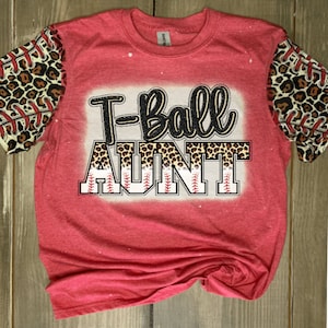 May include: Red t-shirt with a white and black graphic that says "T-Ball Aunt". The graphic has a leopard print design and baseball stitching. The sleeves are leopard print with red baseball stitching.