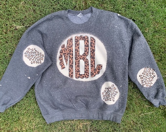 May include: A gray sweatshirt with a white circle on the front featuring the letters "MBL" in a leopard print pattern. The sweatshirt has leopard print circles on the sleeves.