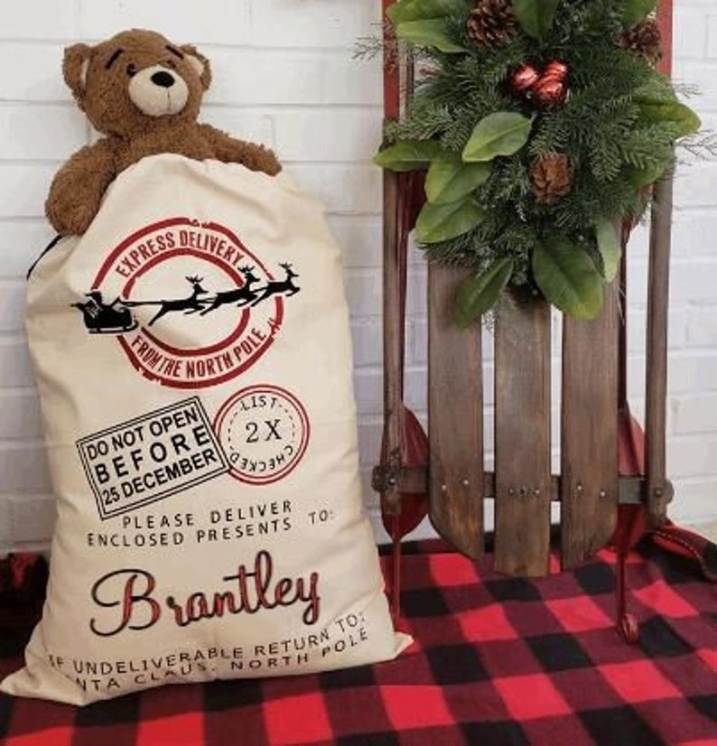 May include: A white canvas Santa sack with red and black lettering. The sack has a red and black stamp that says "List 2X Checked". The sack is decorated with a red and black illustration of Santa's sleigh and reindeer. The text on the sack reads "Express Delivery From The North Pole", "Do Not Open Before 25 December", "Please Deliver Enclosed Presents To Brantley", and "If Undeliverable Return To Santa Claus North Pole".