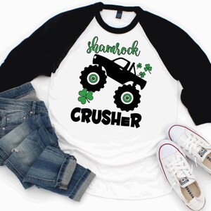 Pu&ograve; includere: Maglietta da baseball bianca e nera con un trifoglio verde e un'immagine di un monster truck nero. Il testo "shamrock crusher" &egrave; stampato sulla maglietta.