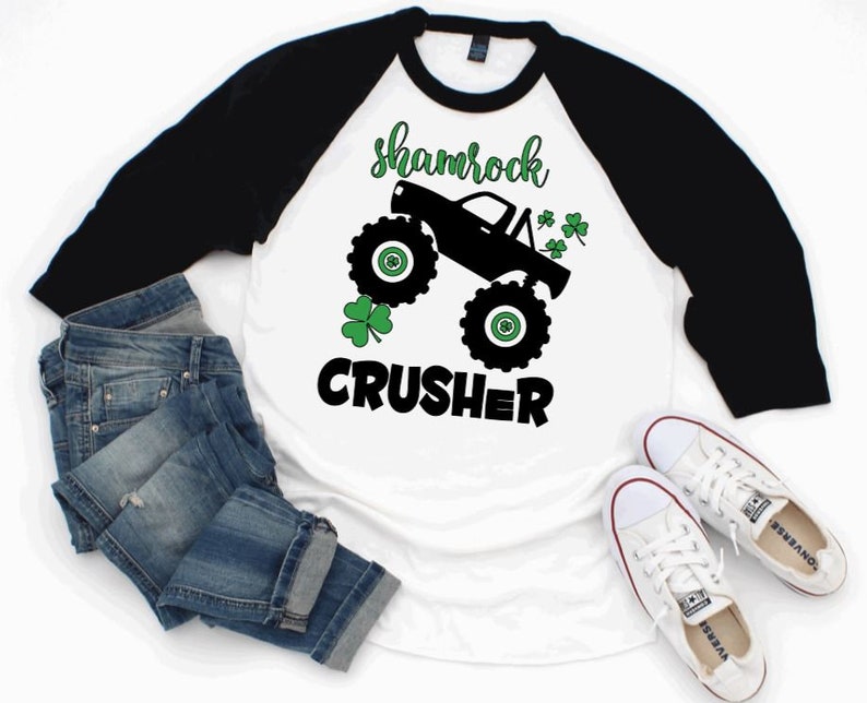 Pu&ograve; includere: Maglietta da baseball bianca e nera con un trifoglio verde e un'immagine di un monster truck nero. Il testo "shamrock crusher" &egrave; stampato sulla maglietta.
