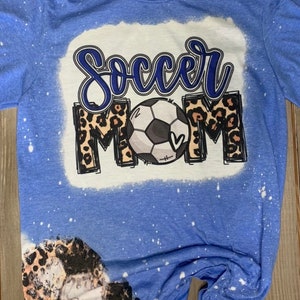 May include: A blue t-shirt with a bleach-splattered design featuring a football and the words "Soccer Mom" in a leopard print.
