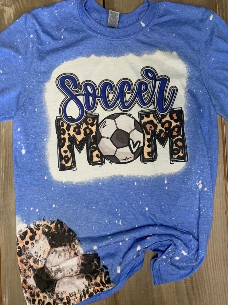 May include: A blue t-shirt with a bleach-splattered design featuring a football and the words "Soccer Mom" in a leopard print.