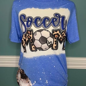 May include: A blue t-shirt with a white bleach design featuring a football and the words "Soccer Mom". The design also includes leopard print accents.