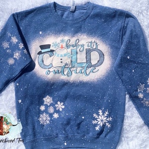 May include: A blue sweatshirt with a white bleach design of a snowman and the text "baby it's cold outside".