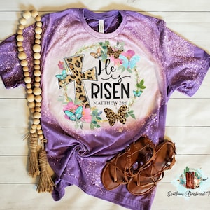May include: Purple bleached t-shirt with a floral wreath design featuring a leopard print cross and the text "He is Risen Matthew 28:6".