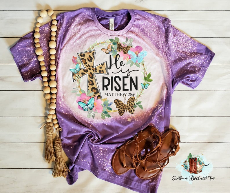 May include: Purple bleached t-shirt with a floral wreath design featuring a leopard print cross and the text "He is Risen Matthew 28:6".