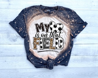 May include: A white t-shirt with a black and white soccer ball design. The shirt has the text "My is on that FIELD" with a leopard print design. The shirt is bleached and has a distressed look.