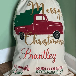 May include: A white canvas Christmas sack with a red truck carrying a green Christmas tree. The sack has the text "Merry Christmas Brantley" in gold and red lettering. It also has a checkbox with the text "nice" checked and the text "naughty" unchecked. The sack has the text "DO NOT OPEN BEFORE DECEMBER 25" in green lettering.