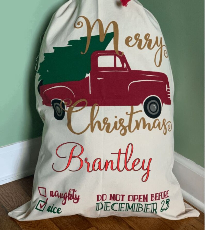May include: A white canvas Christmas sack with a red truck carrying a green Christmas tree. The sack has the text "Merry Christmas Brantley" in gold and red lettering. It also has a checkbox with the text "nice" checked and the text "naughty" unchecked. The sack has the text "DO NOT OPEN BEFORE DECEMBER 25" in green lettering.