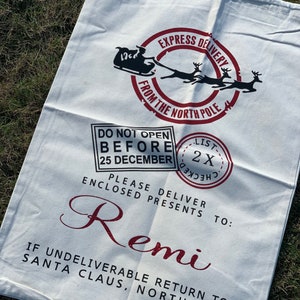 May include: White canvas Santa sack with red and black print. It says "Express Delivery from the North Pole" and "Do Not Open Before 25 December". It also says "Please Deliver Enclosed Presents To: Remi" and "If Undeliverable Return To: Santa Claus, North Pole".