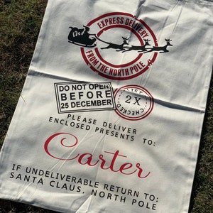 May include: White canvas Santa sack with red text and graphics. The text reads "Express Delivery from the North Pole", "Do Not Open Before 25 December", "Please Deliver Enclosed Presents To: Carter", and "If Undeliverable Return To: Santa Claus, North Pole".