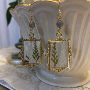 Puede incluir: Pendientes con marco dorado, diseño de helecho prensado y un acento de perla.