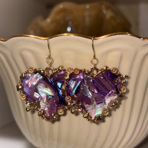 May include: A pair of heart-shaped earrings with iridescent purple and blue abalone shells. The earrings are framed with gold-colored metal and have gold-colored ear wires. The earrings are displayed in a cream-colored bowl.