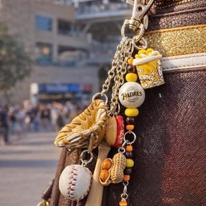 May include: A brown leather bag featuring a metal keychain with miniature baseball-themed charms including a glove, a baseball, a bat, a popcorn bucket, a peanut, and two white beads with the Padres team name and logo, accented by yellow and brown beads.