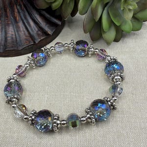 May include: A beaded bracelet with iridescent, faceted beads in shades of blue, purple, and clear. The bracelet features silver-toned metal accents between the beads. The bracelet is on a neutral background.