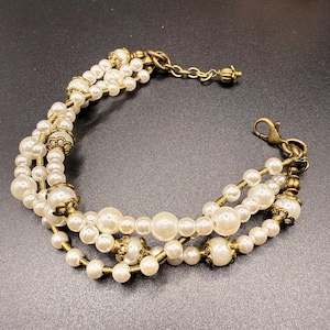 May include: A three-strand bracelet featuring faux pearls in varying sizes. The bracelet has antique gold-colored metal accents and a lobster clasp closure. The pearls are a creamy white color, and the bracelet is designed to be worn on the wrist.