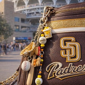 May include: Brown leather purse with gold accents and a San Diego Padres logo. A charm hangs from the zipper with baseball-themed trinkets, including a baseball, glove, and popcorn. The stadium is visible in the background.