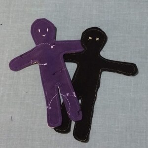 Puede incluir: Dos muñecas de tela, una morada y otra negra, están cosidas juntas. La muñeca morada tiene una cara sonriente y la muñeca negra tiene una cara fruncida.