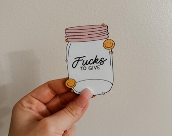 May include: A white sticker with a black outline of a mason jar with a pink lid. The jar has the text "F*cks to Give" written on it. There are small yellow smiley faces and pink stars around the jar.