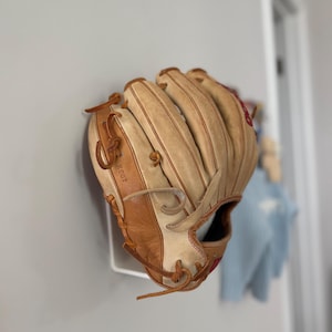 May include: A tan and brown baseball glove with red stitching and leather lacing. The glove is mounted on a white wall hook. The word "KS03" is visible on the glove.