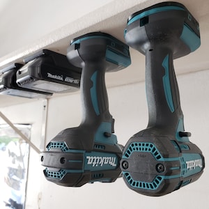 May include: Two blue and black Makita cordless drills hanging from a white shelf. The drills are upside down with the drill bits facing up.